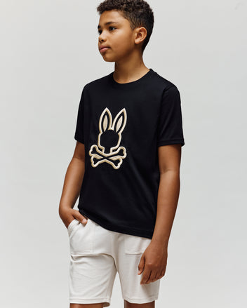 A boy stands with one hand in his pocket, wearing the Psycho Bunny KIDS NICHOLAS EMBROIDERED TEE (KT0200037) and white shorts against a plain light background.