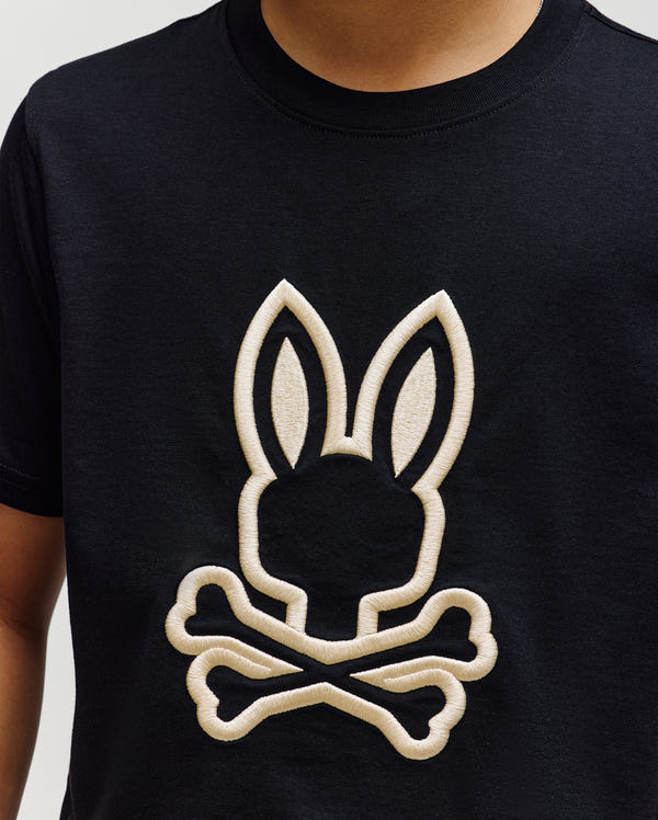 A child wears the Psycho Bunny KIDS NICHOLAS EMBROIDERED TEE (KT0200037), a black t-shirt featuring a large light-colored outline of the Psycho Bunny logo—a bunny head above crossed bones—on the front.