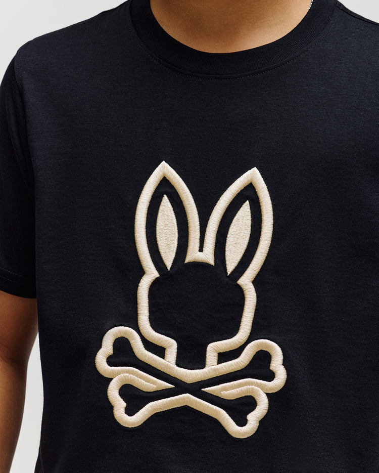 A child wears the Psycho Bunny KIDS NICHOLAS EMBROIDERED TEE (KT0200037), a black t-shirt featuring a large light-colored outline of the Psycho Bunny logo—a bunny head above crossed bones—on the front.