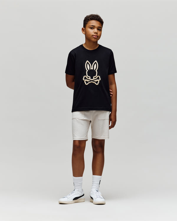 A boy stands against a plain light background, wearing the Psycho Bunny KIDS NICHOLAS EMBROIDERED TEE (KT0200037) in black, paired with light shorts and white sneakers. His arms are relaxed at his sides as he looks slightly right.