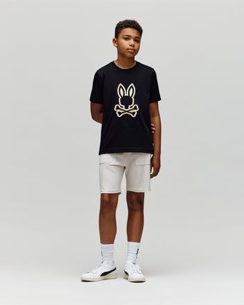 A boy stands against a plain light background, wearing the Psycho Bunny KIDS NICHOLAS EMBROIDERED TEE (KT0200037) in black, paired with light shorts and white sneakers. His arms are relaxed at his sides as he looks slightly right.
