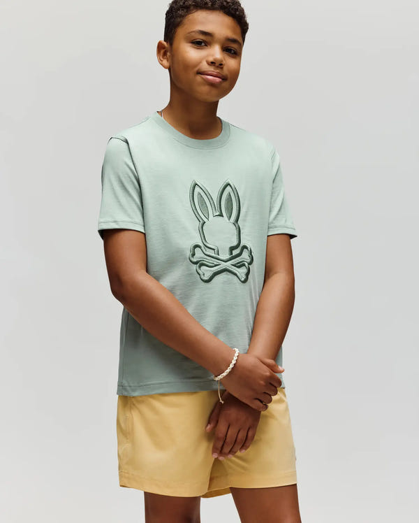 A boy wears the Psycho Bunny KIDS NICHOLAS EMBROIDERED TEE (KT0200037) in light green, featuring a bunny and crossbones design, with light yellow shorts, standing against a plain light background.