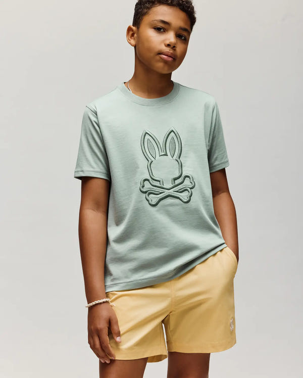 A young person wears the Psycho Bunny KIDS NICHOLAS EMBROIDERED TEE (KT0200037) in light green, featuring a bunny and crossbones design, paired with pale yellow shorts, standing against a plain backdrop with a neutral expression.