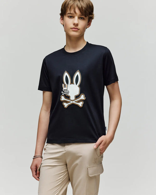 A young person stands against a plain background wearing the Psycho Bunny KIDS WADE GRAPHIC TEE—KT0200054, a black 100% cotton shirt with an embroidered Bunny graphic—and light beige pants with flap pockets.