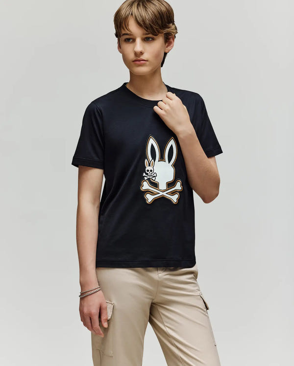 A young person with short brown hair stands against a plain background, wearing the Psycho Bunny KIDS WADE GRAPHIC TEE (KT0200054) with an embroidered Bunny and crossbones design, paired with beige cargo pants. Their left hand rests by their side.