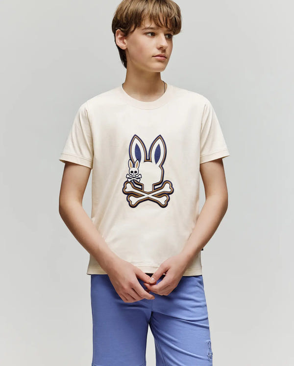 A teenage boy stands against a plain background wearing the Psycho Bunny KIDS WADE GRAPHIC TEE - KT0200054, a 100% cotton shirt with a large embroidered Bunny and crossbones, paired with light blue shorts. He looks to the side, hands clasped.