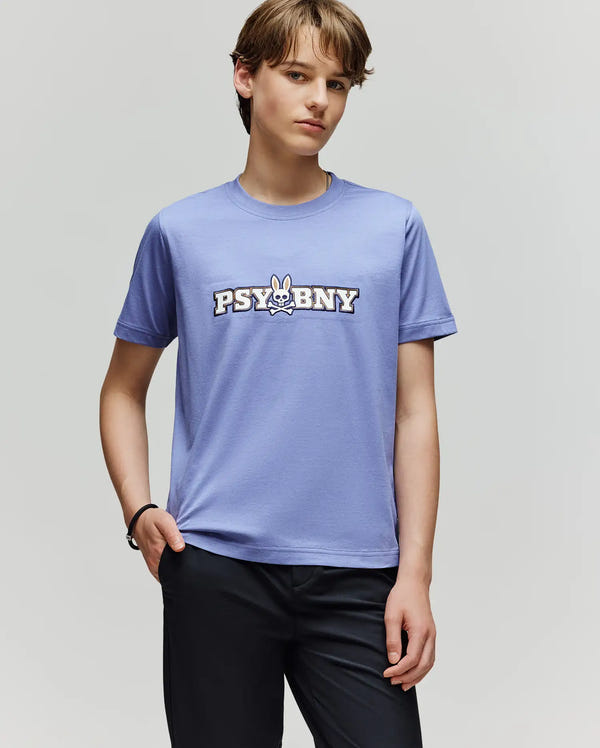 A young person with light brown hair wears a light purple Psycho Bunny KIDS WADE GRAPHIC TEE (KT0200055) featuring "PSYBNY" and a cartoon bunny logo, paired with dark pants, standing against a plain grey background.