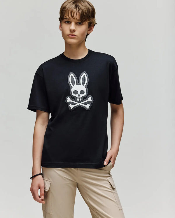 A person with short, light brown hair wears the Psycho Bunny KIDS RYDER OVERSIZED TEE (KT0200056) with an embroidered Bunny skull and crossbones, paired with beige cargo pants, standing against a plain light gray background.