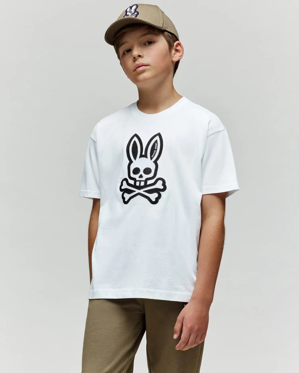 A boy in a beige cap and pants wears the Psycho Bunny KIDS RYDER OVERSIZED TEE (KT0200056), made from soft Pima cotton with an embroidered Bunny skull and crossbones, standing against a plain light gray background.