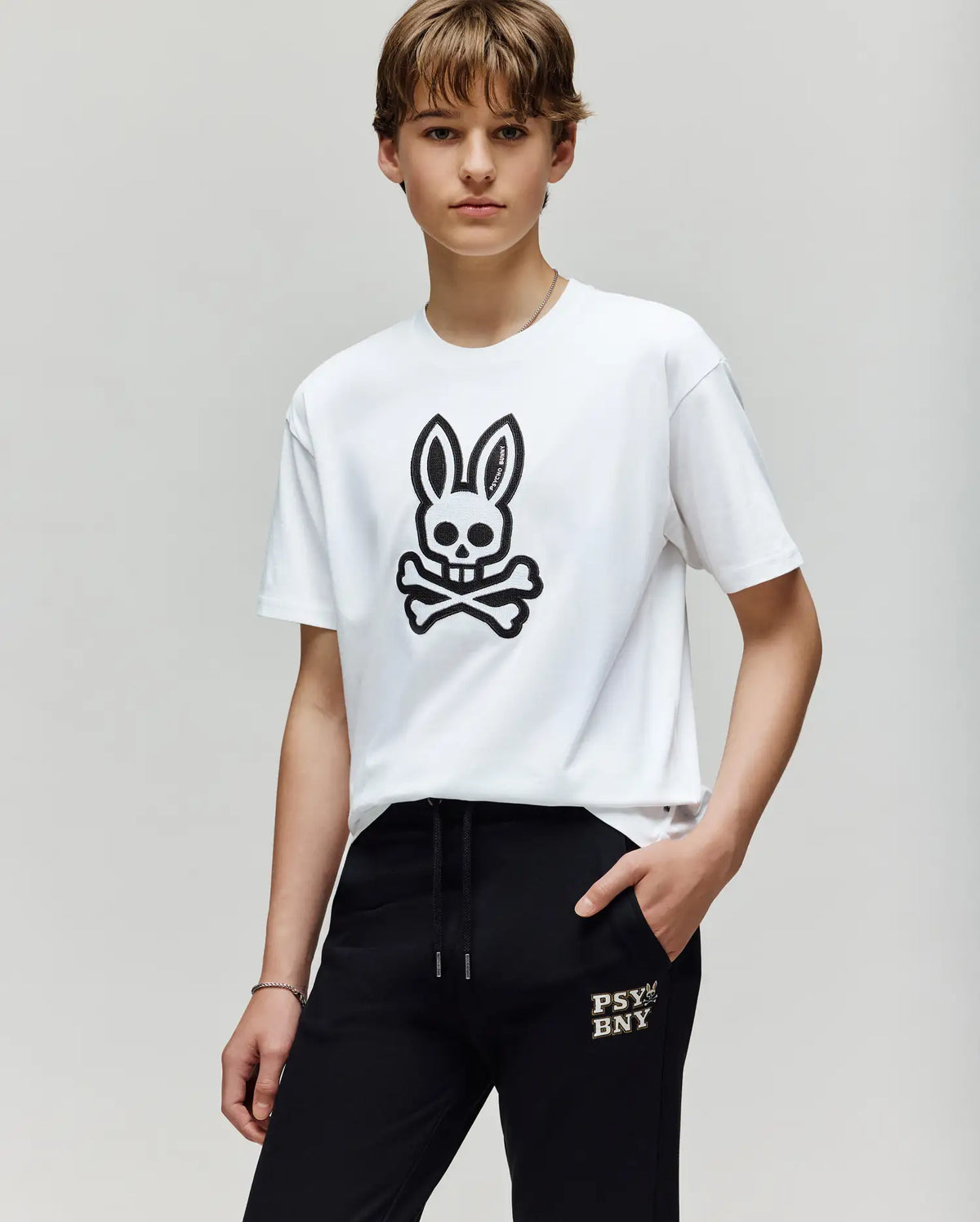 A young person wears the Psycho Bunny KIDS RYDER OVERSIZED TEE (KT0200056), a premium Pima cotton shirt with a large black skull-and-crossbones bunny, paired with black "PSYBNY" pants, standing against a plain light background.