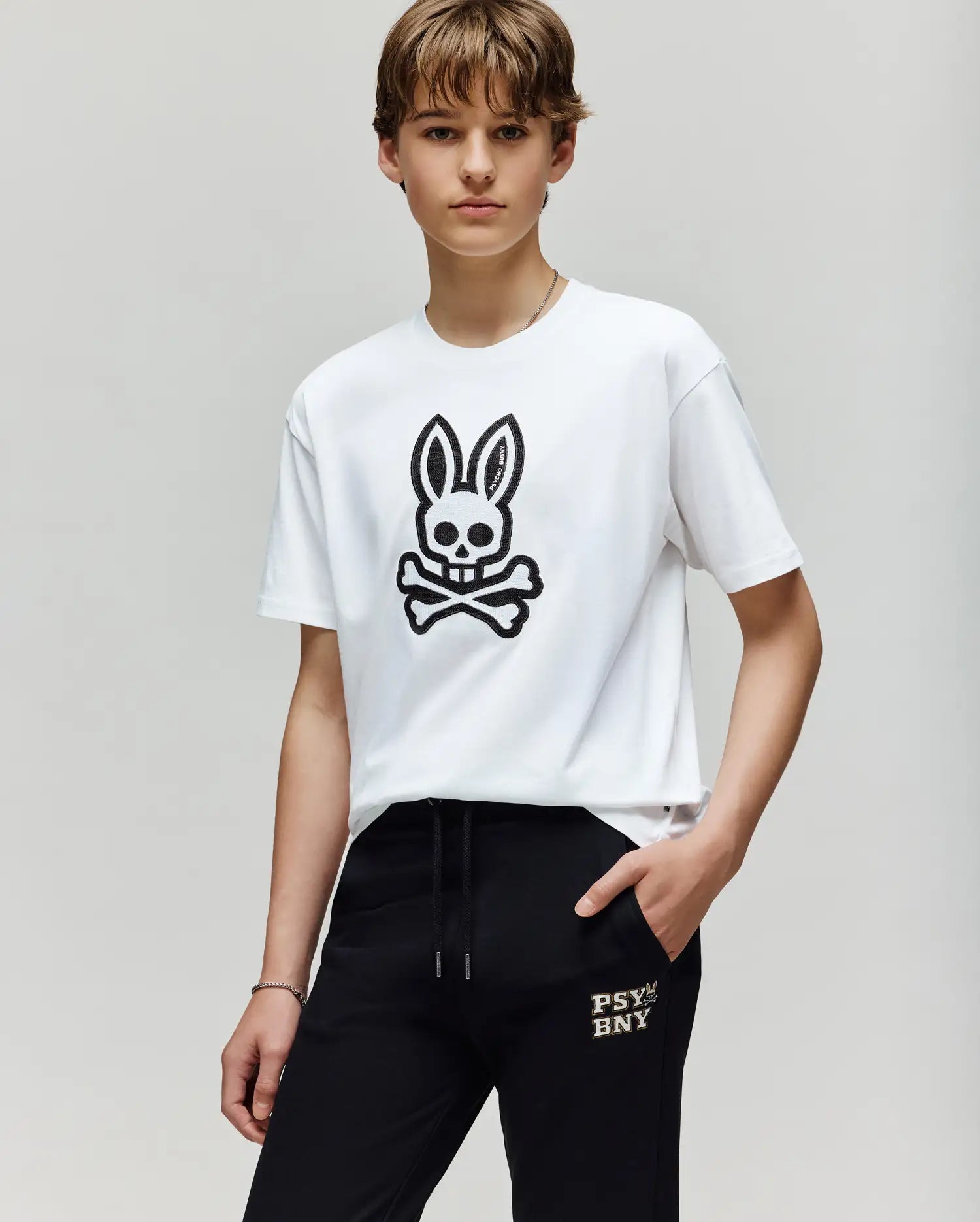 A young person wears the Psycho Bunny KIDS RYDER OVERSIZED TEE (KT0200056), a premium Pima cotton shirt with a large black skull-and-crossbones bunny, paired with black "PSYBNY" pants, standing against a plain light background.