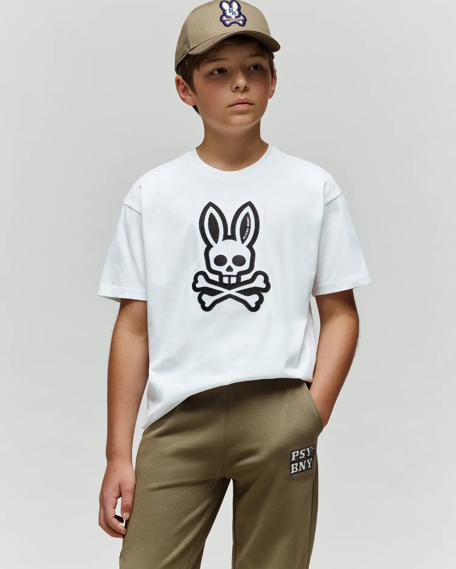 A boy in matching beige cap and pants wears the Psycho Bunny KIDS RYDER OVERSIZED TEE (KT0200056), a white Pima cotton tee with a bold black bunny skull and crossbones logo, standing with one hand in his pocket against a plain background.