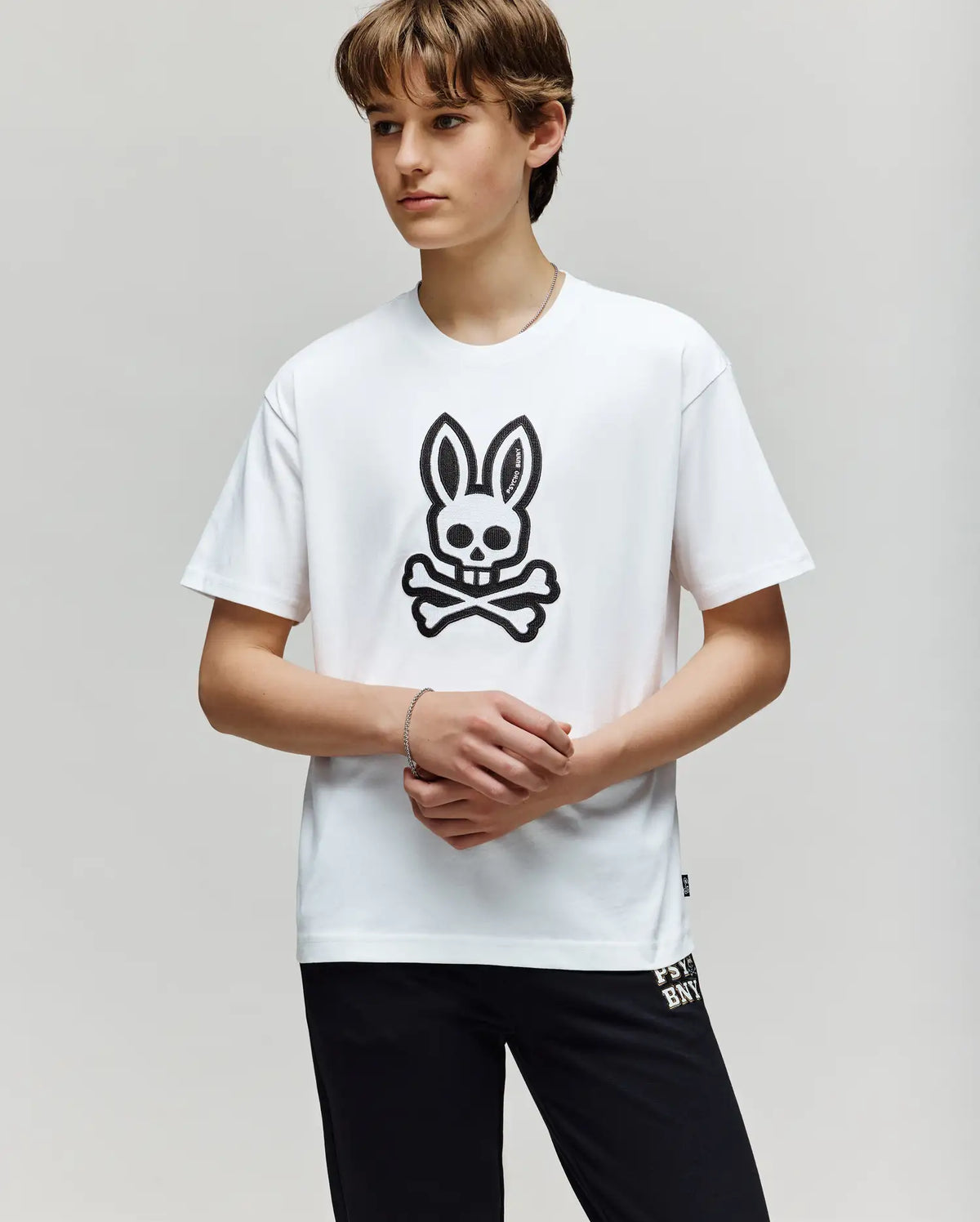 A teen boy with light brown hair wears the Psycho Bunny KIDS RYDER OVERSIZED TEE (KT0200056), featuring an embroidered Bunny skull and crossbones graphic, paired with black pants, against a plain light background.