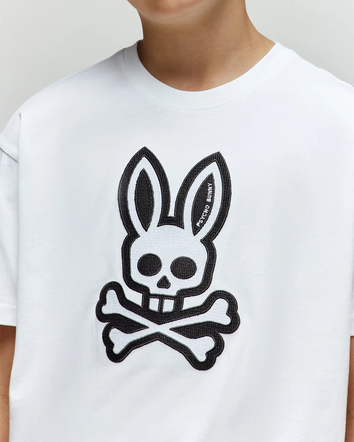 A child wears the KIDS RYDER OVERSIZED TEE (KT0200056) by Psycho Bunny, a soft Pima cotton shirt with a bold black bunny skull-and-crossbones graphic on the front.