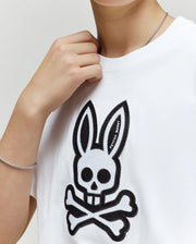 A person wears a silver chain bracelet and necklace with the Psycho Bunny KIDS RYDER OVERSIZED TEE (KT0200056), a premium Pima cotton shirt featuring an embroidered black Bunny skull and crossbones on the chest.