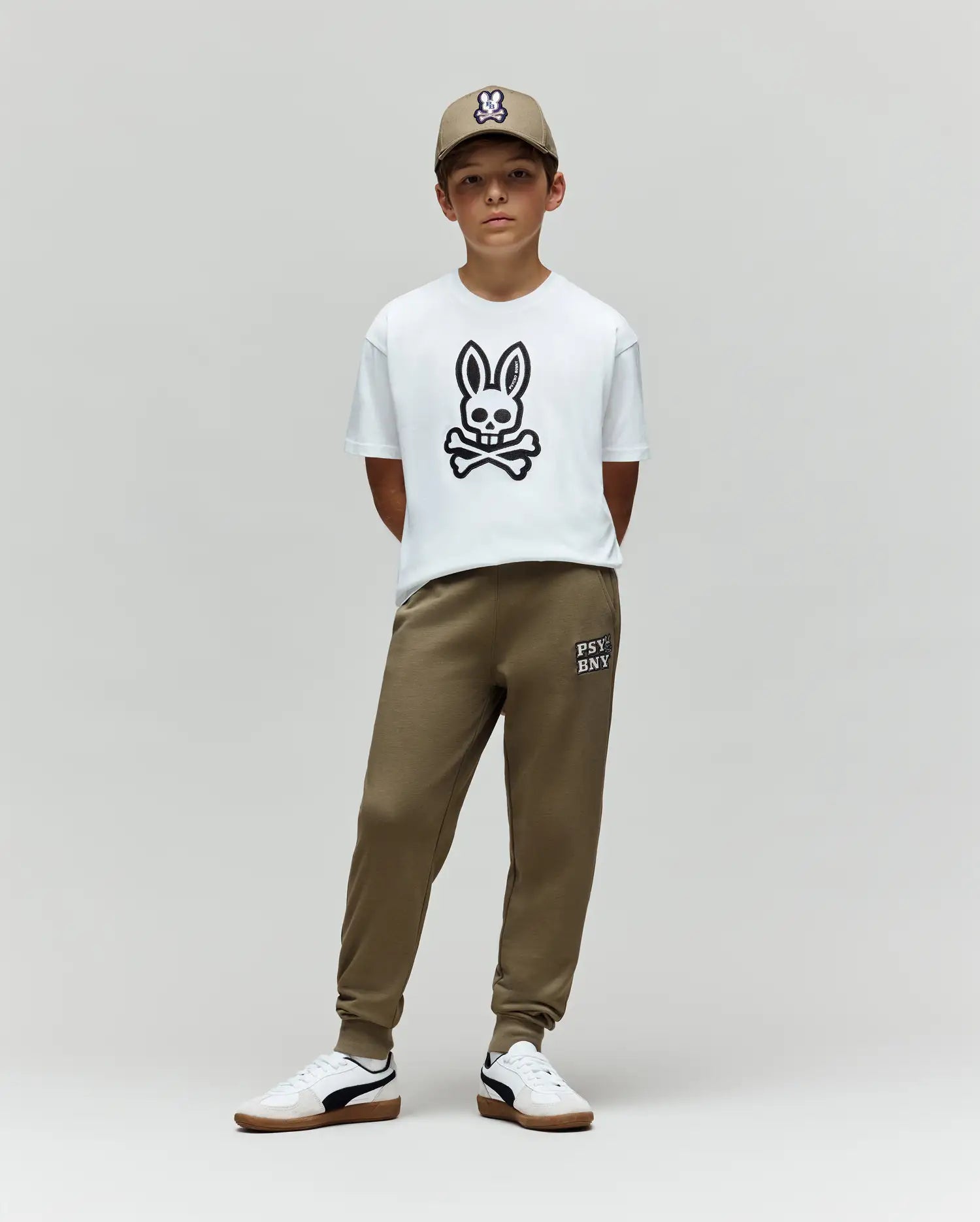 A boy wearing an olive cap, olive jogger pants, white sneakers, and a Psycho Bunny KIDS RYDER OVERSIZED TEE (KT0200056) with a large black embroidered Bunny and crossbones stands against a plain gray background.