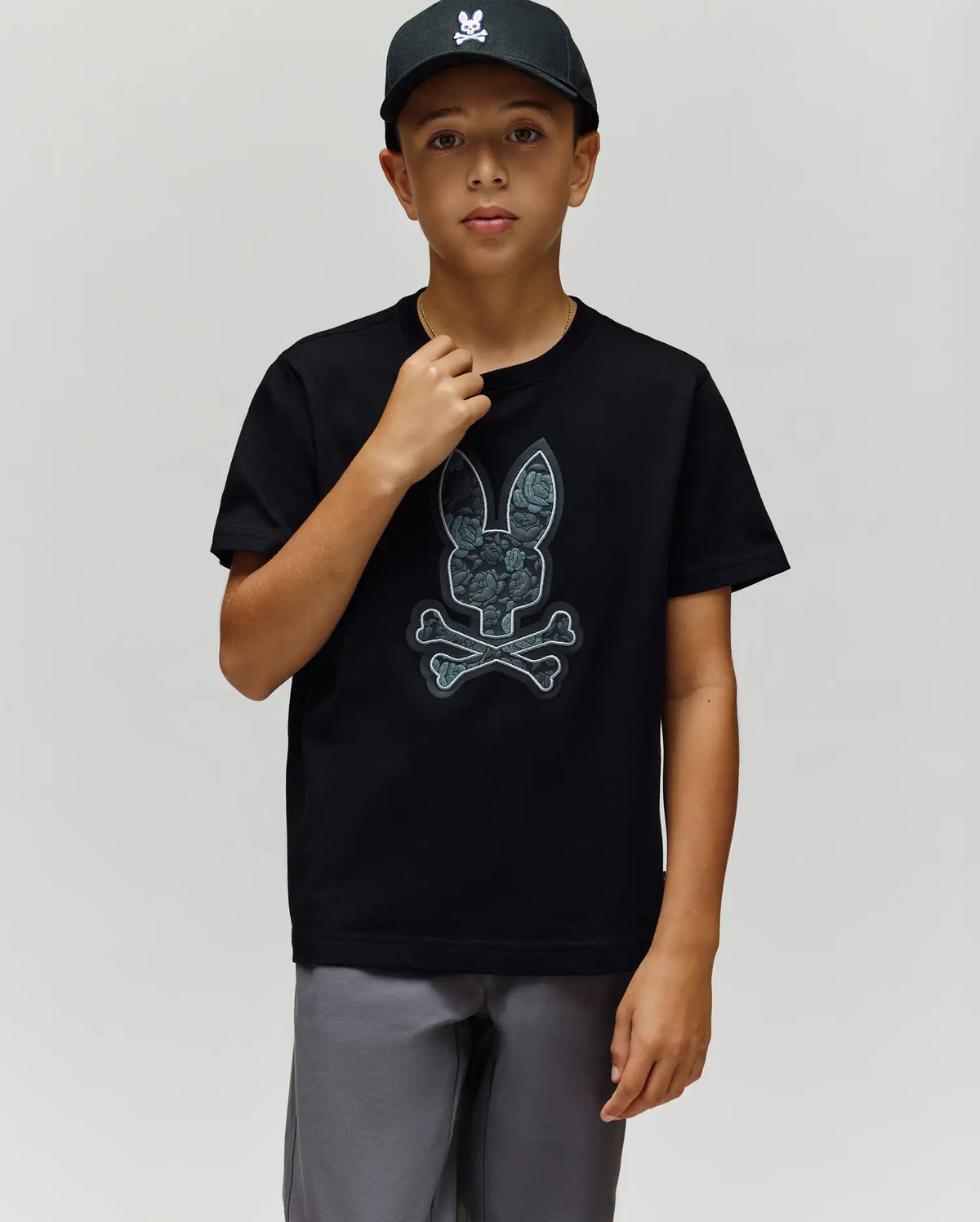 A boy in a black cap and Psycho Bunny KIDS ROWEN BUNNY GRAPHIC TEE (KT0200058) stands against a plain light background. Made in Peru from soft Pima cotton, his tee shows a bunny and crossbones graphic as he looks at the camera.