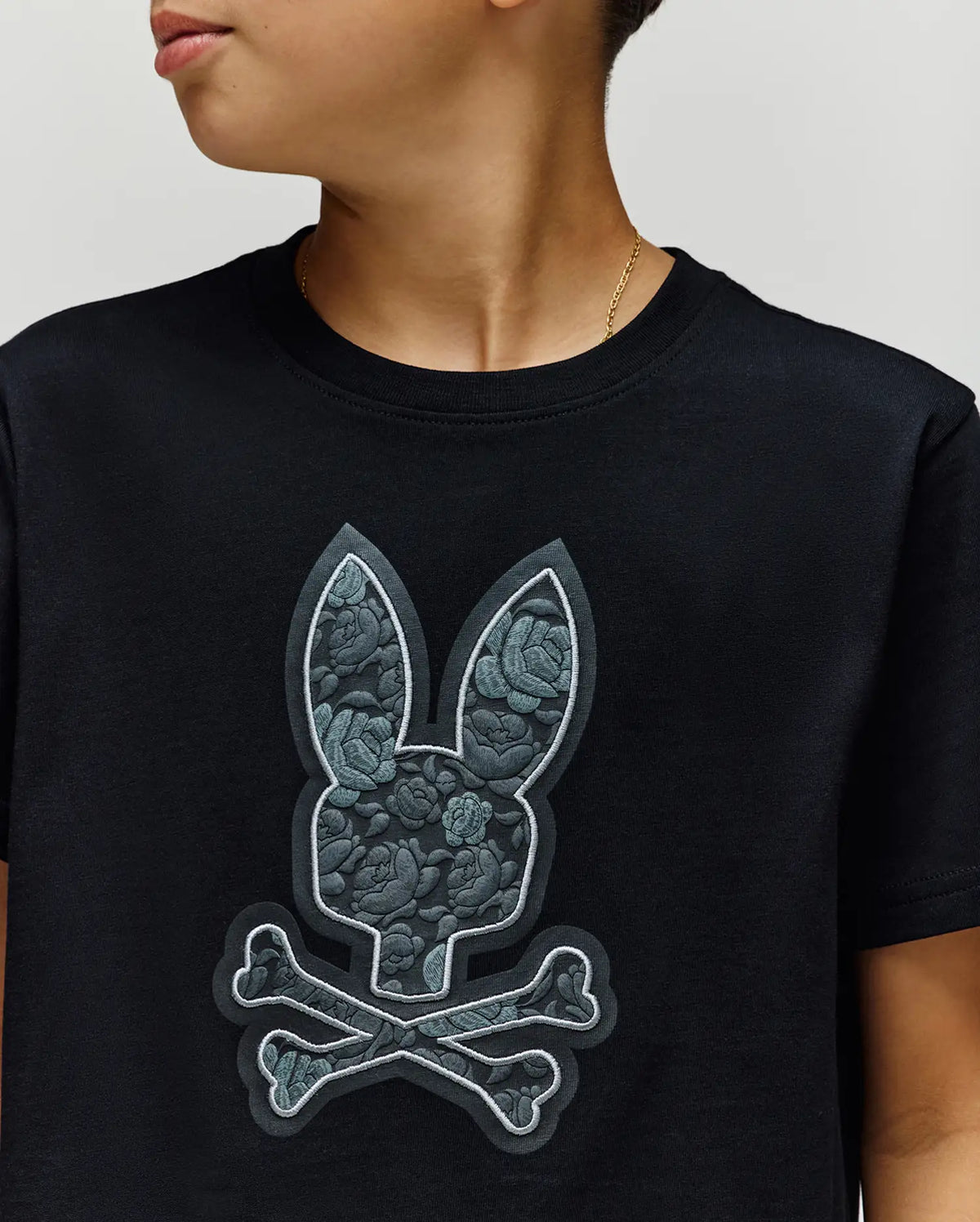 A young person wears the Psycho Bunny KIDS ROWEN BUNNY GRAPHIC TEE - KT0200058, a black Pima cotton shirt made in Peru, featuring a large bunny head and crossbones with a floral texture; only their mouth is visible.