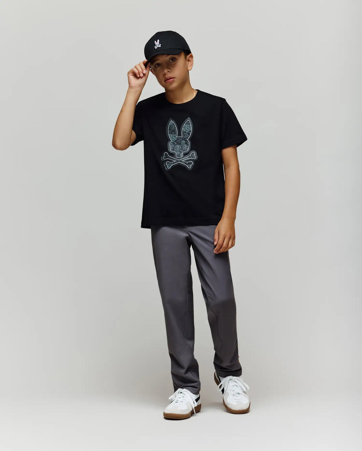 A boy stands against a plain light background, wearing a black cap, gray pants, white sneakers, and the Psycho Bunny KIDS ROWEN BUNNY GRAPHIC TEE (KT0200058), touching the brim of his cap with one hand.