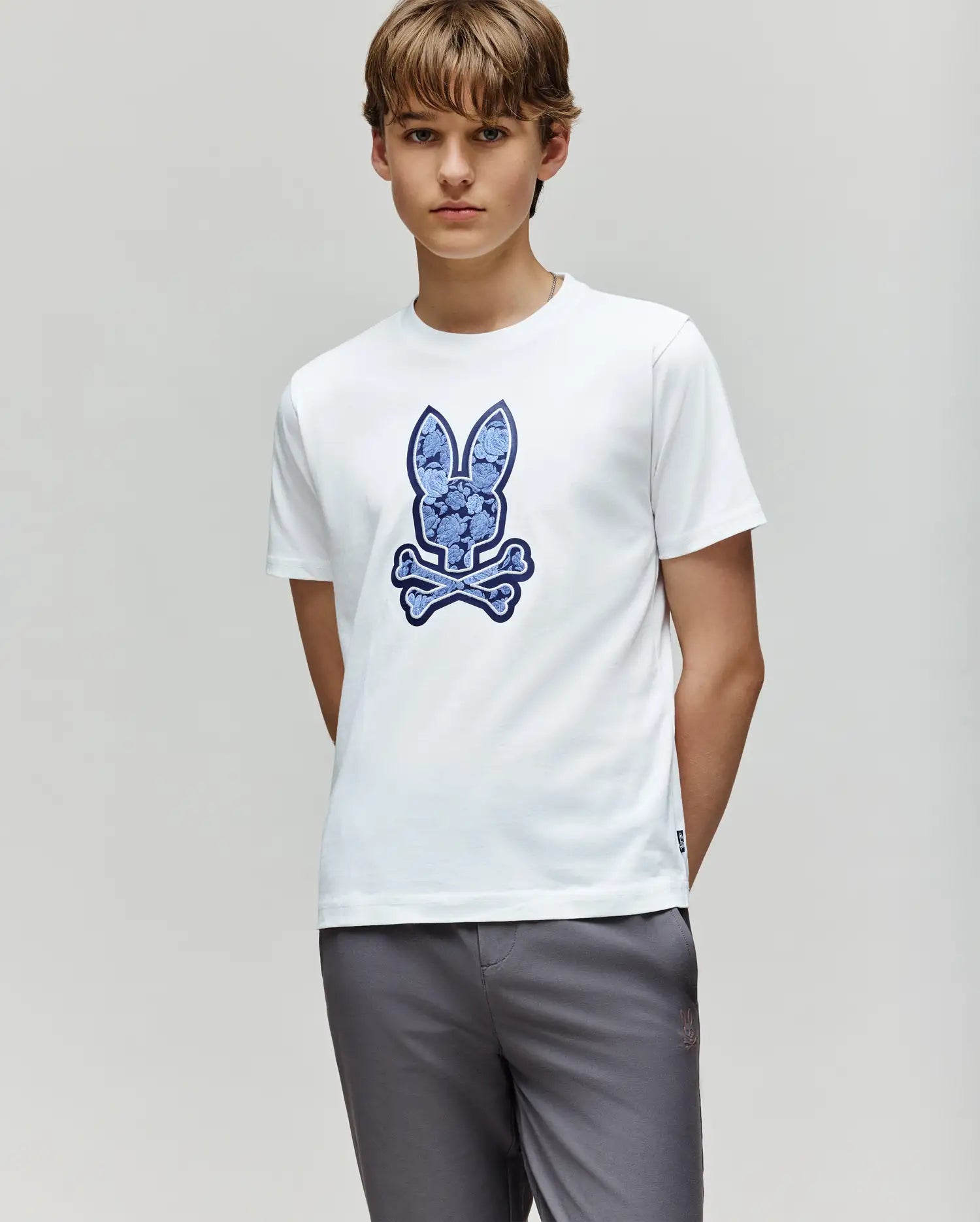 A young person with light brown hair wears the Psycho Bunny KIDS ROWEN BUNNY GRAPHIC TEE (KT0200058), a soft Pima cotton white tee with a large blue bunny and crossbones graphic, paired with light gray pants against a plain background.