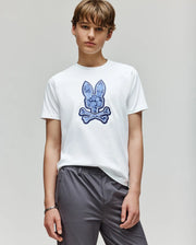 A young person wears the Psycho Bunny KIDS ROWEN BUNNY GRAPHIC TEE (KT0200058), featuring a blue embroidered Bunny and crossbones graphic, paired with gray pants against a plain light background.