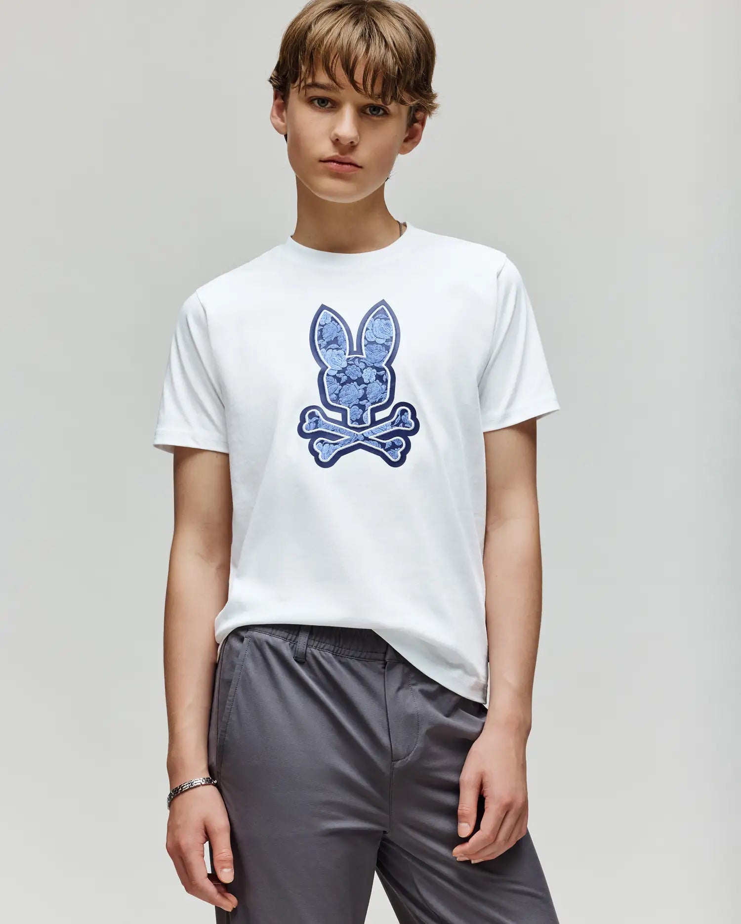 A young person wears the Psycho Bunny KIDS ROWEN BUNNY GRAPHIC TEE (KT0200058), featuring a blue embroidered Bunny and crossbones graphic, paired with gray pants against a plain light background.