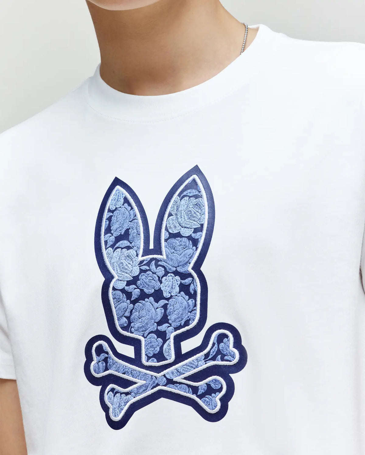 A child wears the Psycho Bunny KIDS ROWEN BUNNY GRAPHIC TEE (KT0200058), a white Pima cotton shirt with a blue floral bunny head and crossbones graphic. Only the lower face and upper body are visible.