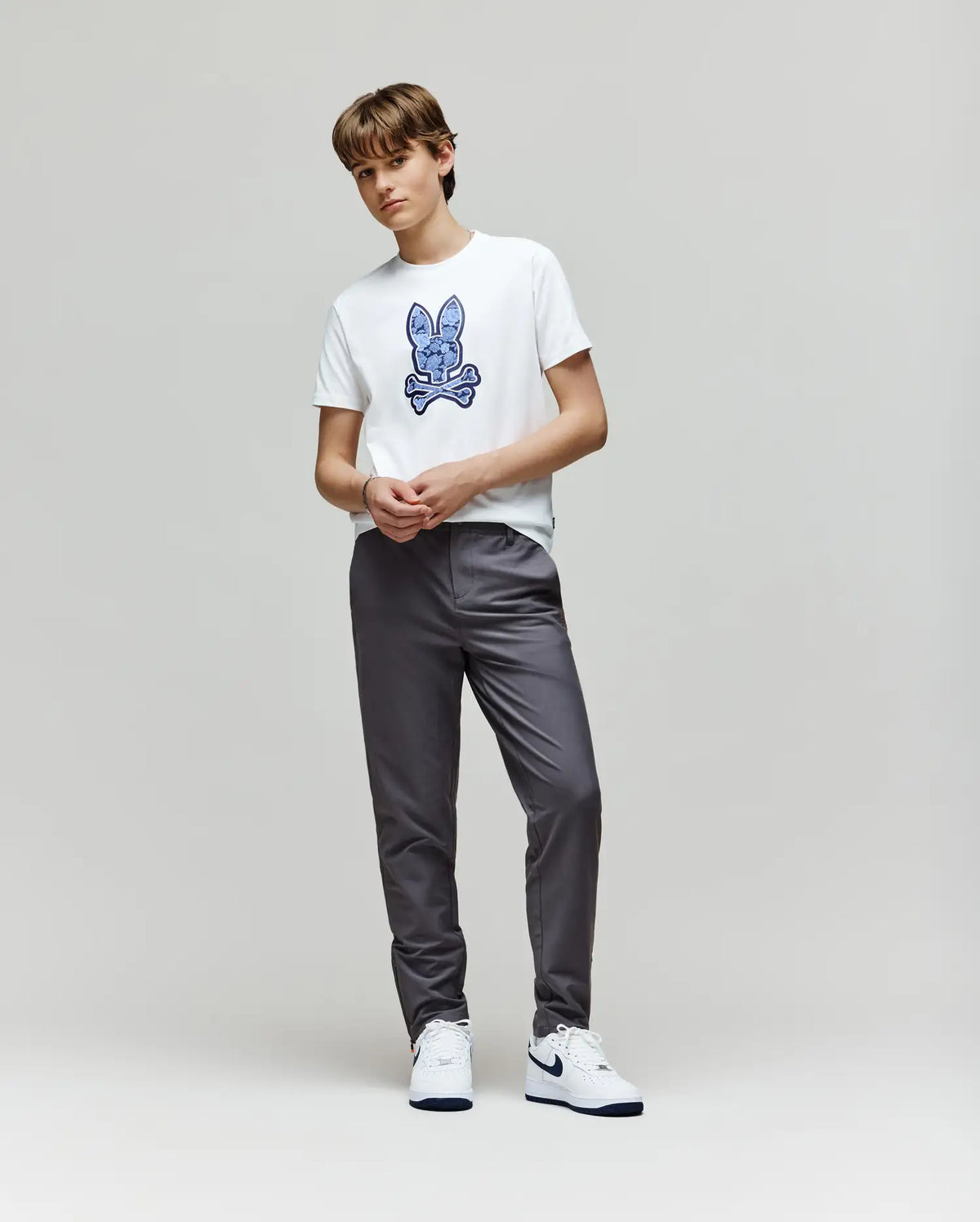 A young person with short hair stands against a plain light background, wearing the Psycho Bunny KIDS ROWEN BUNNY GRAPHIC TEE - KT0200058 with a blue embroidered Bunny graphic, gray pants, and white sneakers, hands together in front.