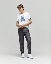 A young person with short hair stands against a plain light background, wearing the Psycho Bunny KIDS ROWEN BUNNY GRAPHIC TEE - KT0200058 with a blue embroidered Bunny graphic, gray pants, and white sneakers, hands together in front.