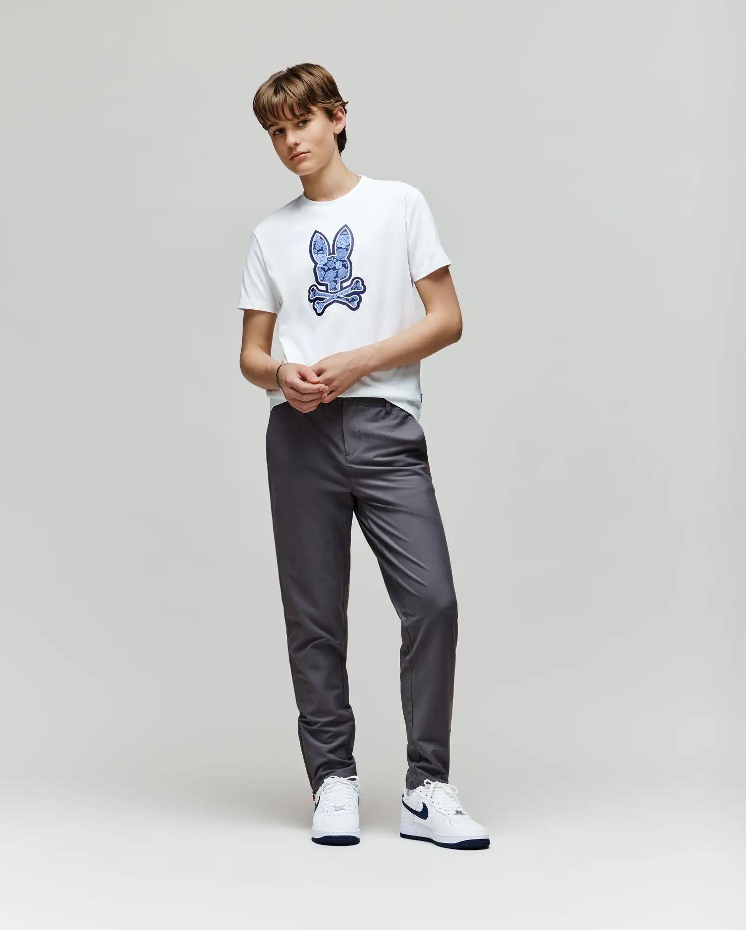 A young person with short hair stands against a plain light background, wearing the Psycho Bunny KIDS ROWEN BUNNY GRAPHIC TEE - KT0200058 with a blue embroidered Bunny graphic, gray pants, and white sneakers, hands together in front.