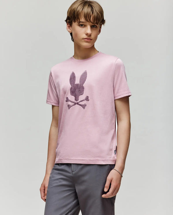 A young person wears the Psycho Bunny KIDS GRANT BUNNY GRAPHIC TEE (KT0200061), a light pink Pima cotton shirt with a stylized bunny and crossbones, paired with gray pants against a plain light background. Made in Peru.