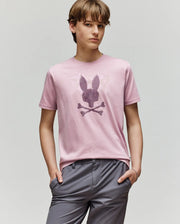 A young person with light brown hair wears Psycho Bunny's KIDS GRANT BUNNY GRAPHIC TEE (KT0200061) in light pink, made from soft Pima cotton, featuring a bunny skull and crossbones, paired with light gray pants against a plain background.