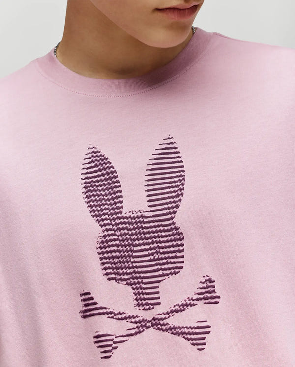 Wearing the Psycho Bunny KIDS GRANT BUNNY GRAPHIC TEE (KT0200061) in light pink, this cropped photo shows the lower face and upper torso. The tee features soft Pima cotton and a striped bunny head above crossed bones graphic.