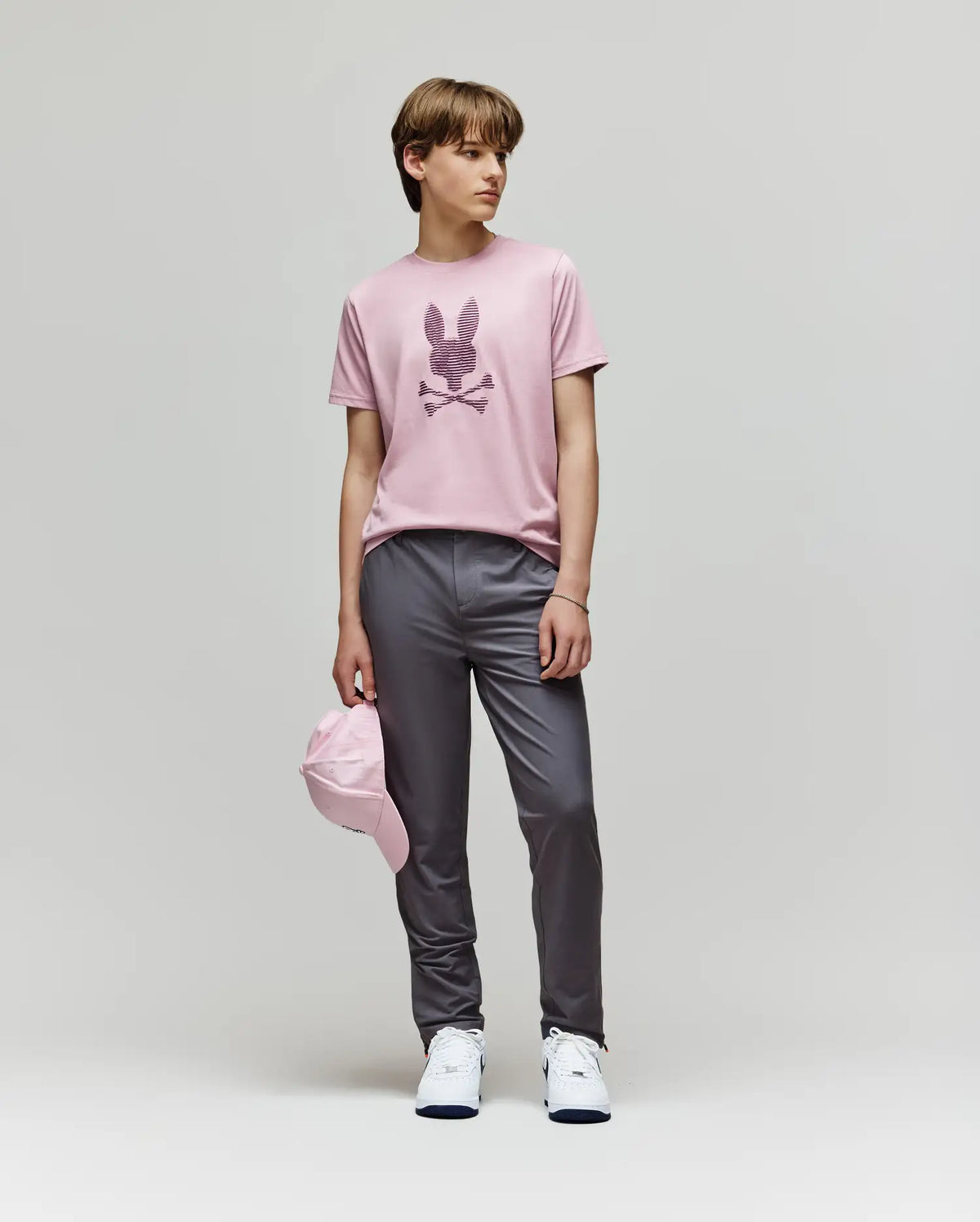 A young person stands against a plain backdrop wearing the Psycho Bunny KIDS GRANT BUNNY GRAPHIC TEE (KT0200061), paired with gray pants, white sneakers, and holding a pink cap at their side.