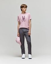A young person stands against a plain backdrop wearing the Psycho Bunny KIDS GRANT BUNNY GRAPHIC TEE (KT0200061), paired with gray pants, white sneakers, and holding a pink cap at their side.