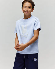 A smiling boy wears the Psycho Bunny KIDS GRANT BUNNY GRAPHIC TEE (KT0200061) in light blue Pima cotton with an HD Bunny and crossbones print, paired with navy shorts featuring an embroidered patch, against a light gray background.