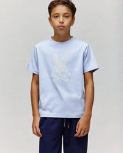 A boy with short brown hair wears the Psycho Bunny KIDS GRANT BUNNY GRAPHIC TEE (KT0200061), featuring an HD-printed Bunny and crossbones design, along with navy blue pants, standing against a plain background.