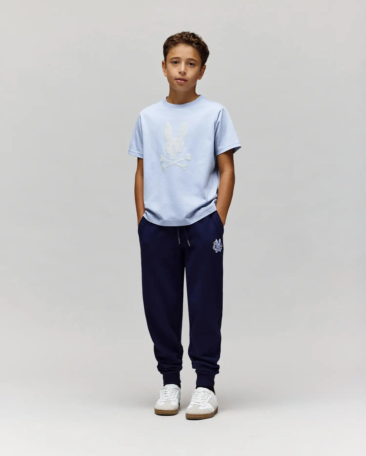 A boy stands against a light gray background, wearing the Psycho Bunny KIDS GRANT BUNNY GRAPHIC TEE (KT0200061), navy jogger pants, and white sneakers. His hands are in his pockets as he looks straight ahead.