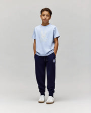 A boy stands against a light gray background, wearing the Psycho Bunny KIDS GRANT BUNNY GRAPHIC TEE (KT0200061), navy jogger pants, and white sneakers. His hands are in his pockets as he looks straight ahead.