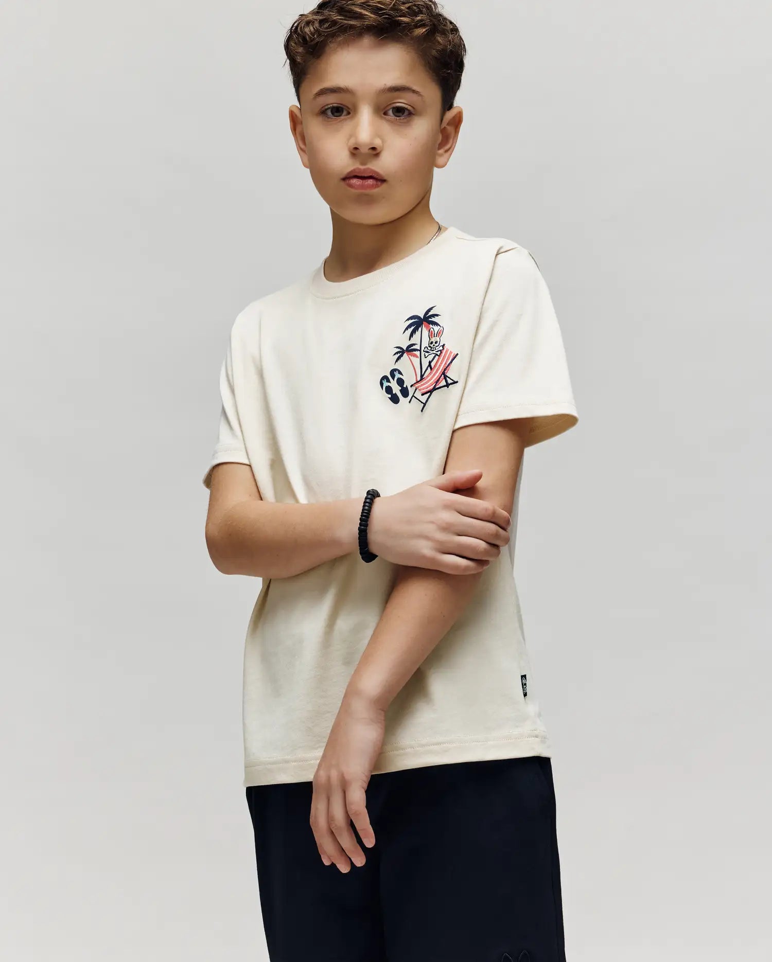 A boy with short curly hair wears the Psycho Bunny KIDS SONNY GRAPHIC TEE (KT0200091), a cream Pima cotton t-shirt with an embroidered palm tree and surfboard graphic, paired with black shorts, standing against a light background.