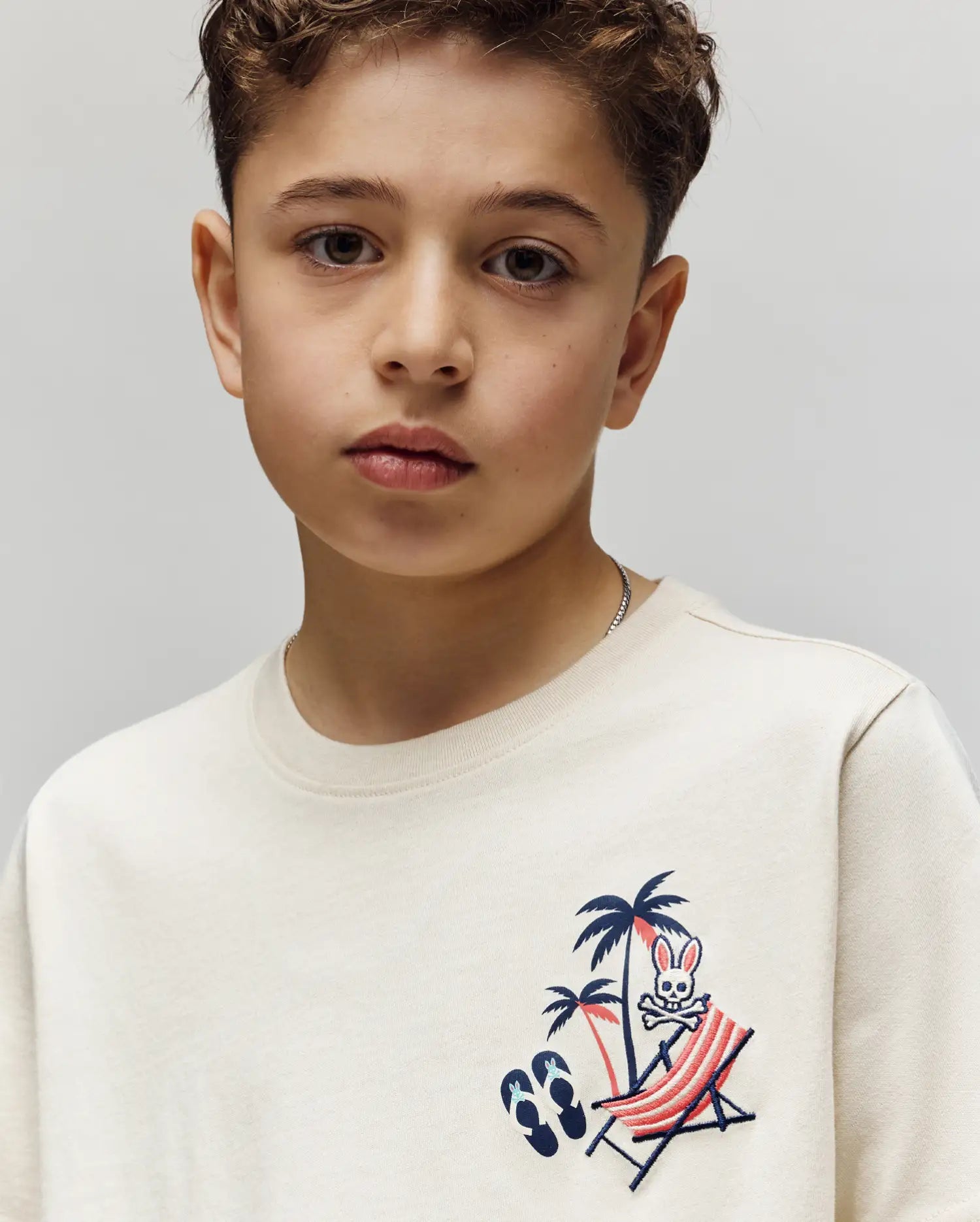 A young boy with short curly hair wears the Psycho Bunny KIDS SONNY GRAPHIC TEE (KT0200091), a cream Pima cotton shirt featuring an embroidered bunny lounging on a striped deck chair under a palm tree. The background is plain and light.