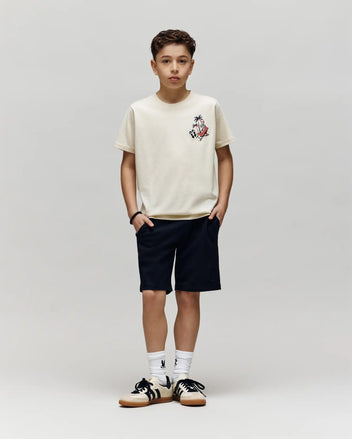 A boy stands against a gray background, wearing the Psycho Bunny KIDS SONNY GRAPHIC TEE (KT0200091) in light beige with an embroidered chest graphic, paired with dark shorts, white socks, and black-and-white sneakers. His hands are in his pockets.