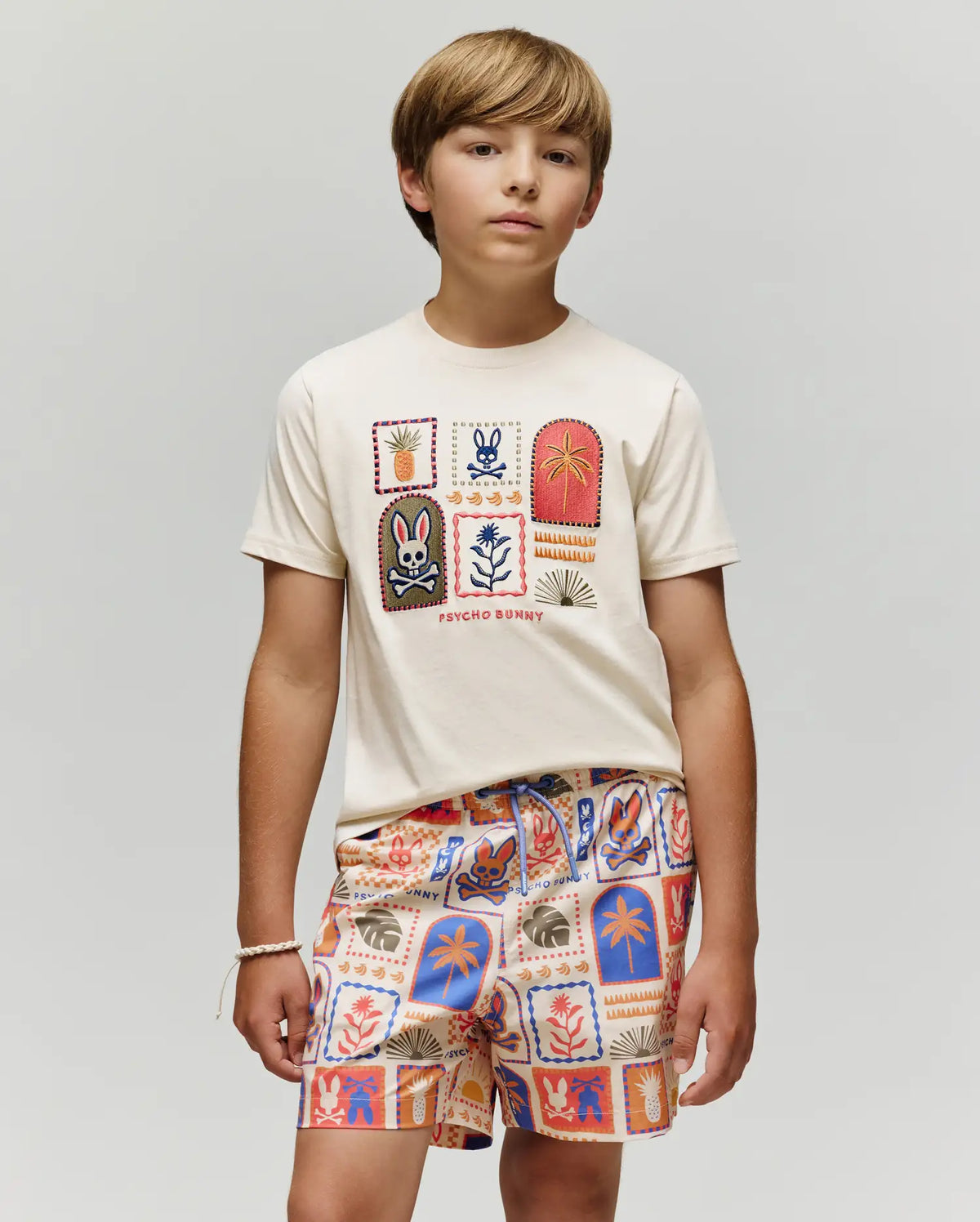 A boy with light brown hair wears the Psycho Bunny KIDS SUNDEW GRAPHIC TEE (KT0200092), a cream-colored Pima cotton T-shirt with colorful graphic patches, paired with matching Beach Bunny print shorts, standing calmly against a light gray background.