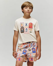 A boy with light brown hair wears the Psycho Bunny KIDS SUNDEW GRAPHIC TEE (KT0200092), a cream-colored Pima cotton T-shirt with colorful graphic patches, paired with matching Beach Bunny print shorts, standing calmly against a light gray background.
