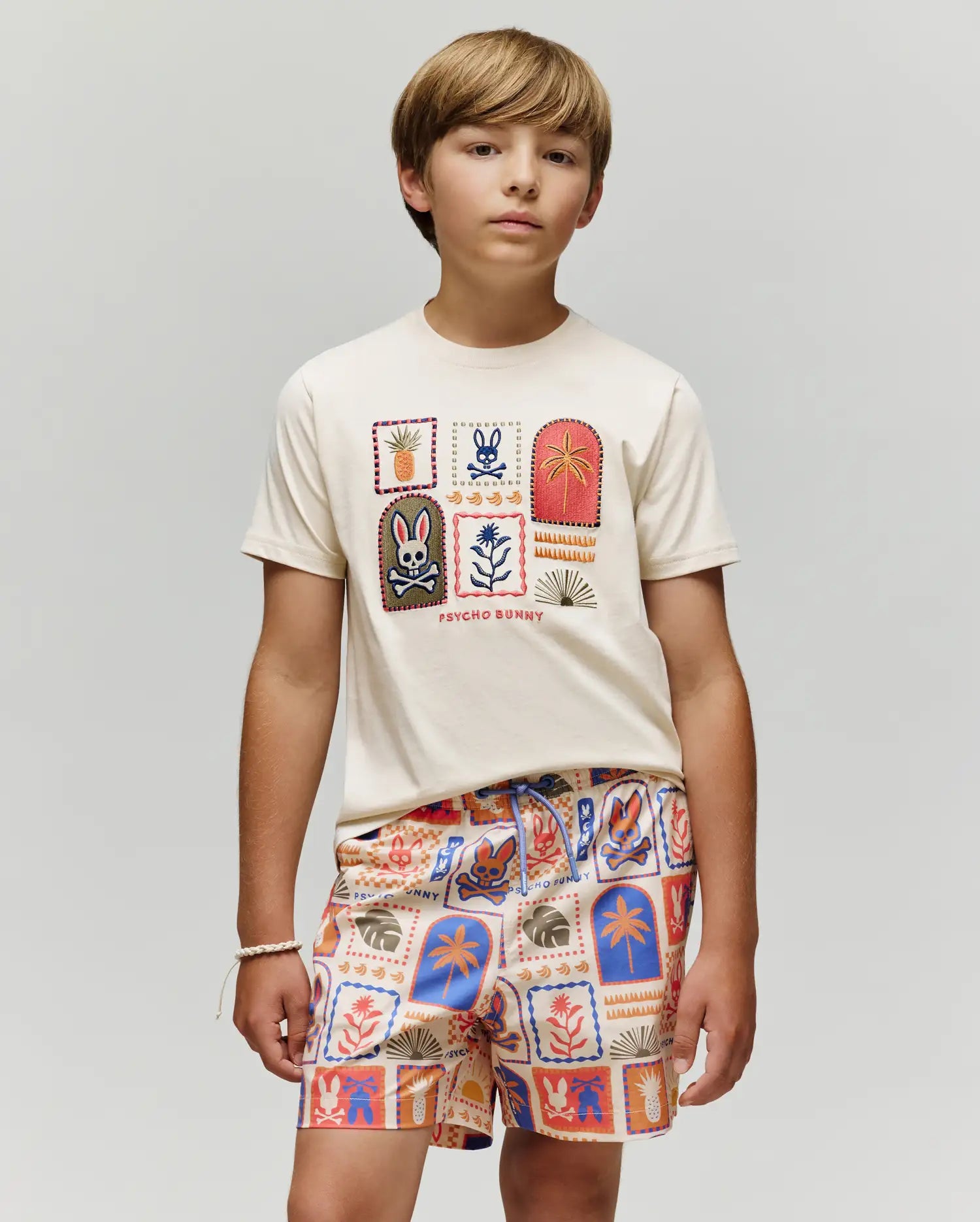 A boy with light brown hair wears the Psycho Bunny KIDS SUNDEW GRAPHIC TEE (KT0200092), a cream-colored Pima cotton T-shirt with colorful graphic patches, paired with matching Beach Bunny print shorts, standing calmly against a light gray background.