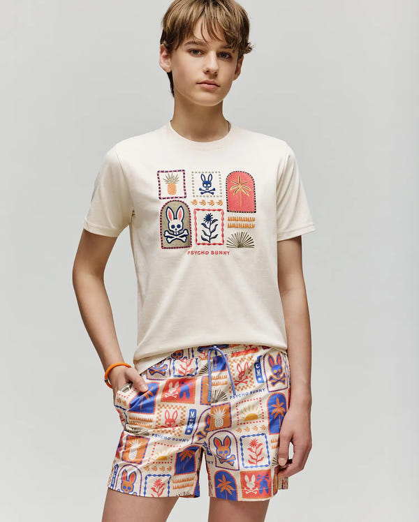 A teenager with light brown hair wears the Psycho Bunny KIDS SUNDEW GRAPHIC TEE - KT0200092, a cream Pima cotton shirt with playful graphics, paired with orange, blue, and cream patterned shorts against a plain light background.