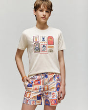 A teenager with light brown hair wears the Psycho Bunny KIDS SUNDEW GRAPHIC TEE - KT0200092, a cream Pima cotton shirt with playful graphics, paired with orange, blue, and cream patterned shorts against a plain light background.