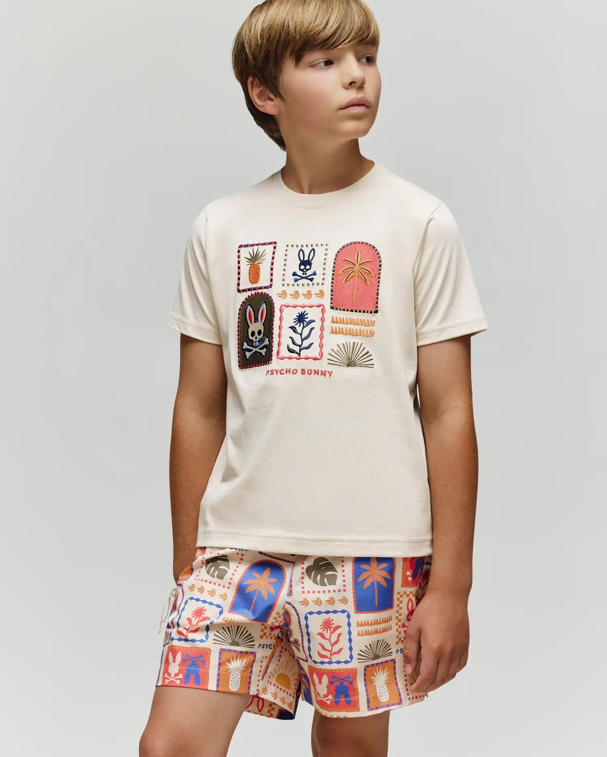 A boy wearing the Psycho Bunny KIDS SUNDEW GRAPHIC TEE (KT0200092), a cream Pima cotton shirt with colorful graphic patches, and matching Beach Bunny print shorts stands against a plain light gray background.
