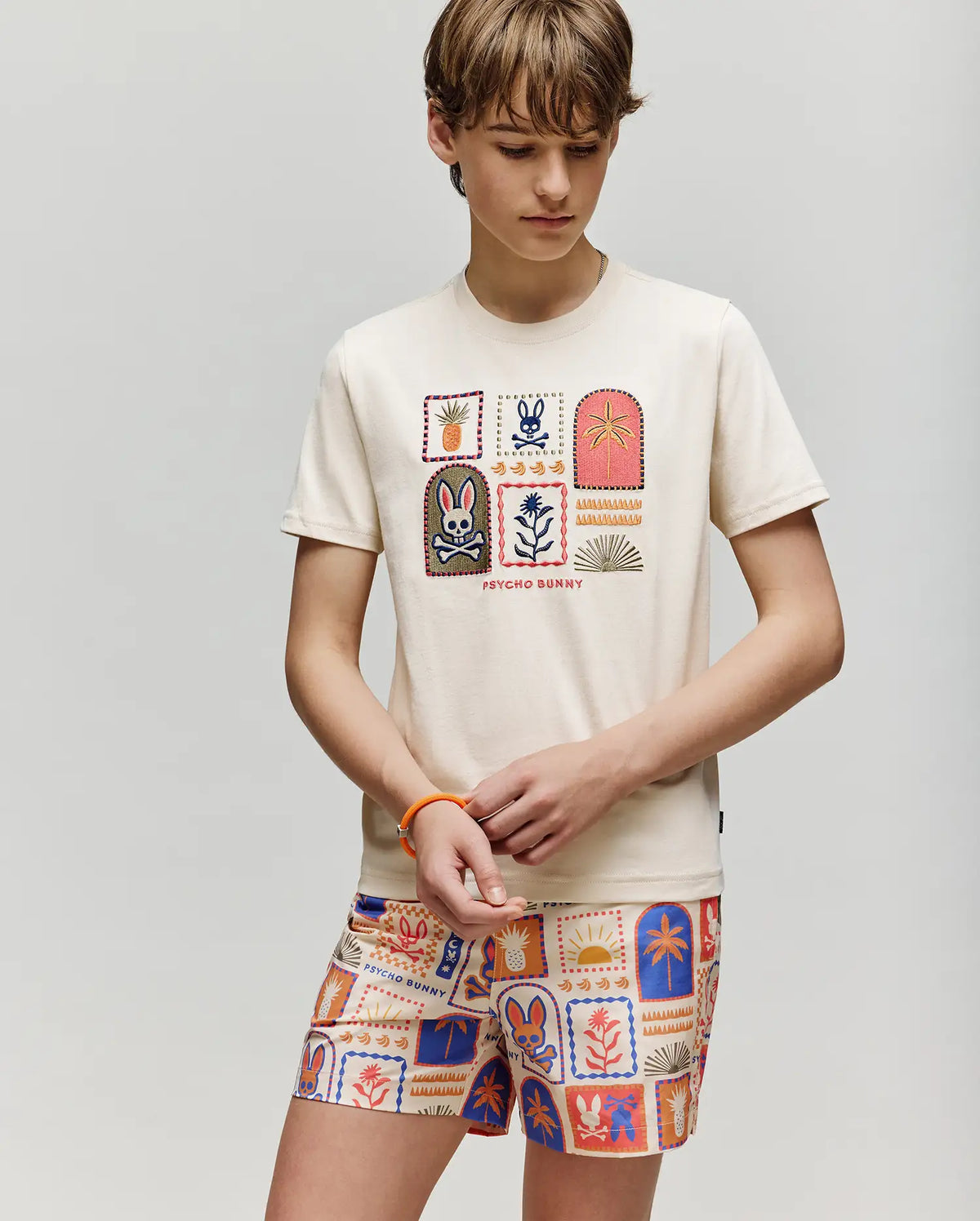 A child models the Psycho Bunny KIDS SUNDEW GRAPHIC TEE (KT0200092), a cream-colored tee with playful Beach Bunny graphics, paired with colorful shorts, standing against a plain light background, looking down with hands clasped.