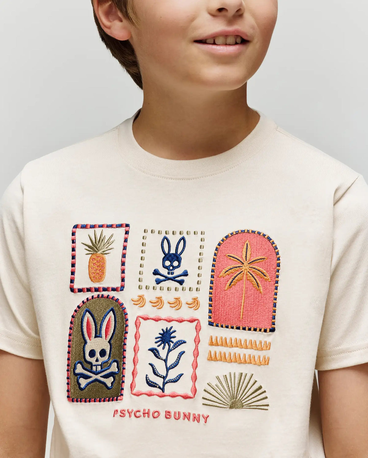 A child wears the Psycho Bunny KIDS SUNDEW GRAPHIC TEE (KT0200092), a cream Pima cotton shirt featuring fun embroidered bunnies, skull, pineapple, palm tree, plant, and shapes in a playful Beach Bunny print with "PSYCHO BUNNY" at the hem.